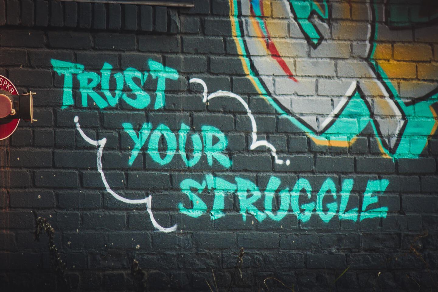 turquoise graffiti sprayed on a darkish-blue colored brick wall reading: trust your struggle. another graffiti is partially visible looking like the letter q